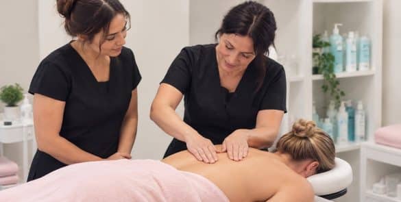 Illustration of a tutor demonstrating an upper-back massage while a learner observes in a training room.