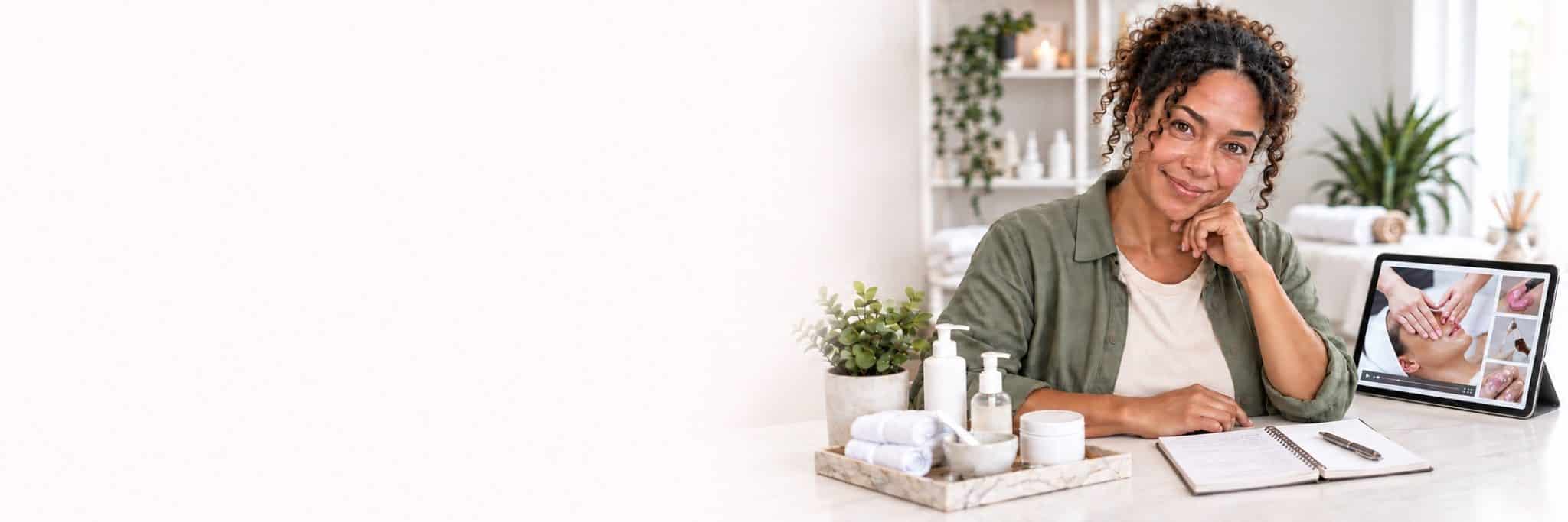 Beauty learner with an iPad showing a facial treatment tutorial, an open notepad and skincare products.