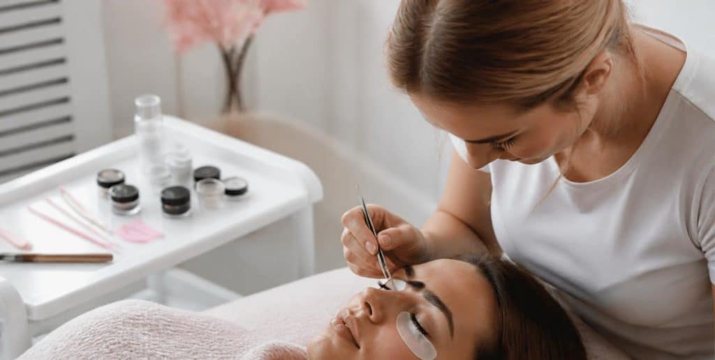 A lash and brow technician applying individual lashes with fine tweezers, with tint pots and a lash lift kit on the tray beside her