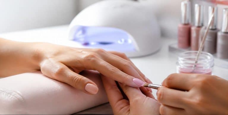A nail technician working on a client’s gel manicure at a salon desk, with a UV lamp and gel polishes alongside