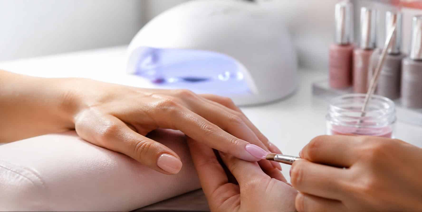 A nail technician working on a client’s gel manicure at a salon desk, with a UV lamp and gel polishes alongside